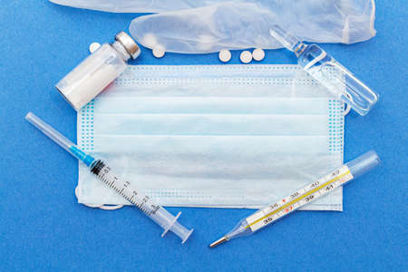 medical protective masks with thermometer, syringe, ampoule, medicine bottle, pills on a blue...