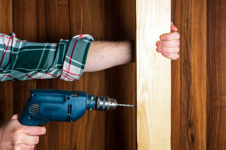 Woodworker drills a hole with an electric drill in a wood board. Close up of a foreman hand at work. Working environment in a carpentry workshopの写真素材