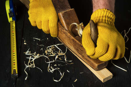 Woodworker using a hand plane to clean up a wooden board. Hands of the master in gloves close up at work. Working environment in carpentry workshopの写真素材
