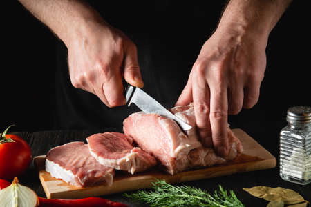 Chef Butcher cutting pork meat with knife on kitchen, cooking food. Vegetables and spices on the kitchen table to prepare a delicious lunchの写真素材