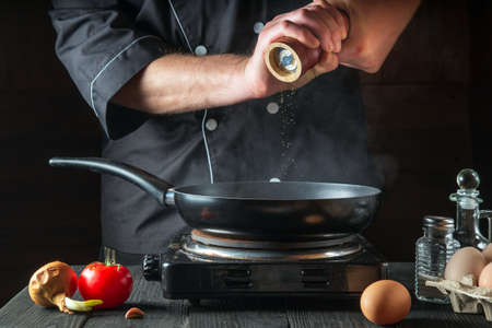The chef adds pepper while cooking eggs in a pan. Work environment on vintage kitchen tableの写真素材