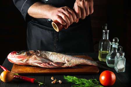 The cook prepares fresh fish bighead carp sprinkling pepper. Preparing to cook fish food. Working environment in the restaurant or cafe kitchenの写真素材