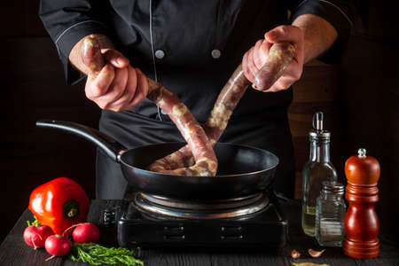The chef holds the raw meat sausage before cooking. Working environment in a restaurant kitchen with vegetables and spices on the vintage tableの写真素材