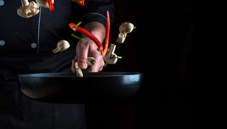 Hand of a professional chef tosses pieces of vegetables and mushrooms on a frying pan on a black...