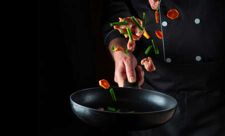 The hand of a professional chef tosses pieces of vegetables and beef meat on a frying pan on a black...