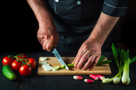 Chef is hands close-up cuts young green onions on a restaurant kitchen cutting board for salad. Vegetable diet or snack idea.の写真素材