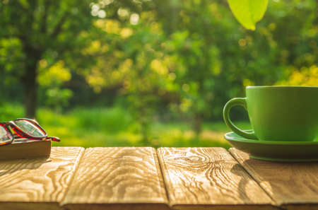 A cup of hot coffee and glasses with a book on a wooden table in a green garden in the morning. Summer background.の写真素材