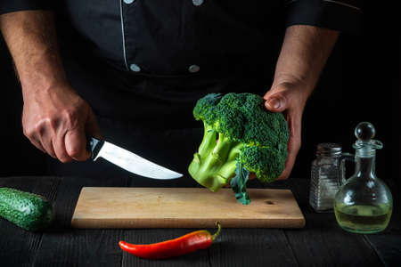 Professional chef cuts fresh Broccoli with a knife for salad on vintage kitchen table with fresh vegetables. Cooking and restaurant or cafe conceptの写真素材