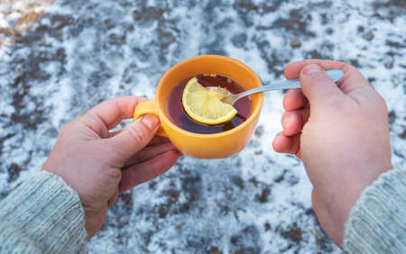 People hands hold an orange mug of hot herbal tea in cold winter. Prevention of viral diseases lemon tea. Spoon in person is handの写真素材