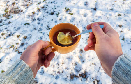 Person hand hold an orange mug of hot herbal tea in cold winter. Prevention of viral diseases lemon tea. Spoon in person is hand.の写真素材