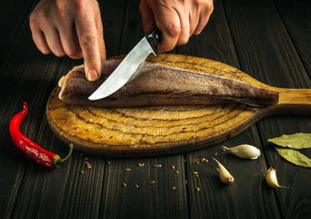 Professional chef cuts fresh Merluccius fish on a wooden cutting board with a knife. Home cooking of a delicious fish dish according to an old recipeの写真素材