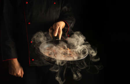 Professional chef prepares food in a frying pan with steam on black background. The concept of restaurant and hotel serviceの写真素材