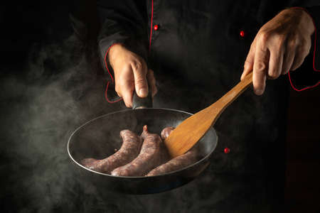 The chef is frying sausages for hot dogs in a frying pan. Close-up of a cook hands with a hot pan in...