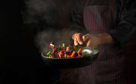 The hands of a professional chef throws pieces of vegetables into a hot frying pan with steam on a black background. Hotel cooking concept.の写真素材