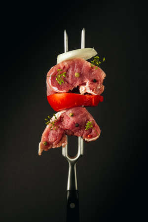 Raw veal meat with spices, dill, onion and tomato on a fork before grilling or barbecue. Hotel menu concept on black backgroundの写真素材