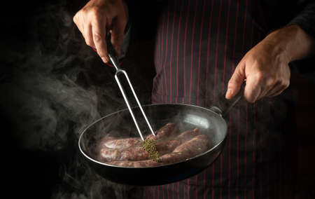 The chef fries sausages for hot dogs in a frying pan. Close-up of the cook's hands with a hot pan...