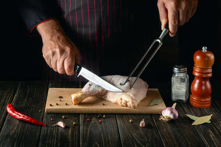 The chef cuts a fresh raw chicken drumstick on a dark background. Nearby on the kitchen table are ingredients for cooking and spices.の写真素材