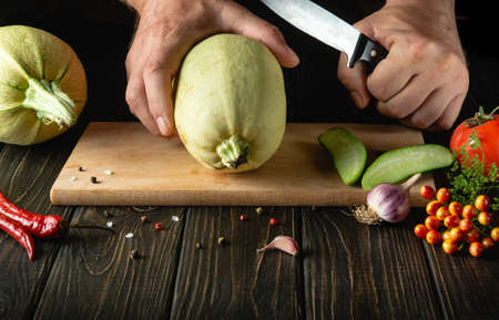 Slicing vegetable marrow with a knife before cooking by the hands of the cook on a wooden cutting board. In cooking, the thick flesh of zucchini is used.の写真素材