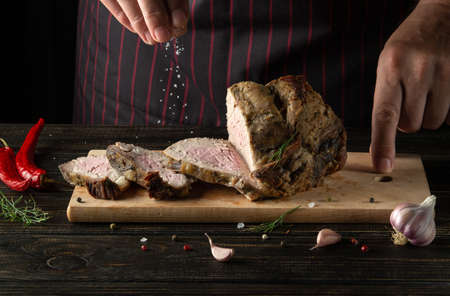 The cook salts the fried and sliced marbled beef steak. Served on a rustic wooden board. Work environment on dark kitchen table with spices and condimentsの写真素材