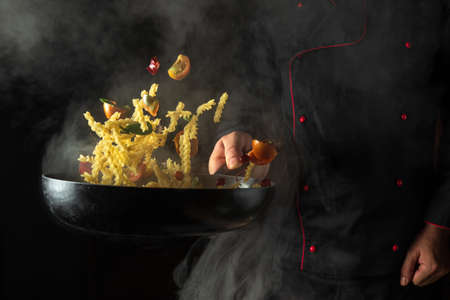 Cook prepares food in a hot pan with steam on a black background. The concept of restaurant and hotel service. Thai food pasta with spices and vegetablesの写真素材