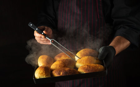 The baker bakes pies or buns on a hot baking sheet taken out of the oven. Checking readiness with a fork in the chef's hand. Black space for menu or recipe.の写真素材