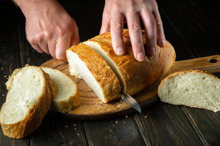 The hands of a cook with a knife cut fresh wheat bread before breakfast or lunch. The concept of...