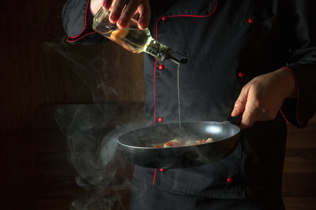 The chef adds olive oil to fried carrots and onions in a frying pan. Cooking vegetables for lunch. Restaurant or hotel menu on a dark background.の写真素材