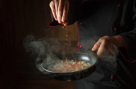 The chef adds spices to fried carrots with onions in a frying pan. Cooking vegetables for lunch. Restaurant or hotel menu on a dark background.の写真素材