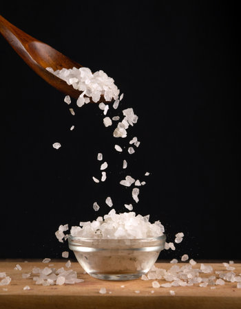 Large white salt is poured from a wooden spoon into a glass salt cellar. Copy space on a black...