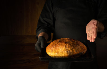 The chef or baker presents in his hand a sheet pan with freshly baked wheat bread. Free dark space...