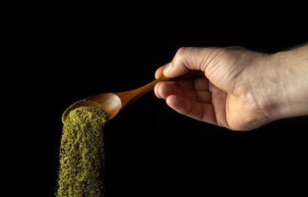 Mixture of dry chopped herbs is poured from a wooden spoon to add to a medicinal drink or tea. Free space for advertising on a black backgroundの写真素材