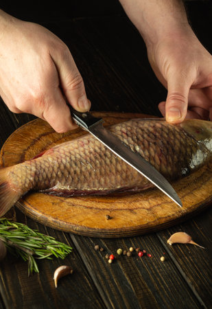 Close-up of the chef's hands with a knife on the kitchen table before cleaning the carp. Cooking a fish dish on the kitchen table with spices and pepper.の写真素材