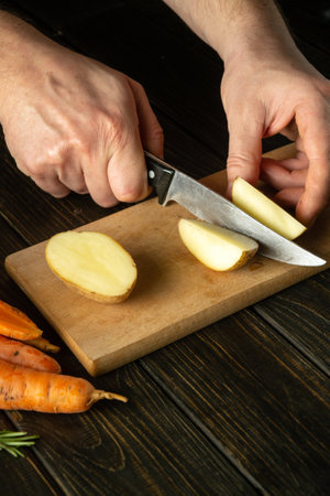 The chef cuts fresh potatoes on a wooden cutting board. Close-up of cook hands while preparing vegetarian food with vegetables.の写真素材