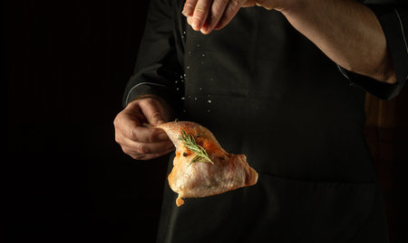The cook adds salt to a raw chicken leg. The concept of cooking a meat dish for lunch on a black background.の写真素材