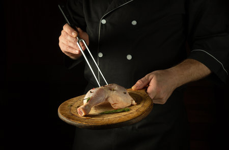 The chef prepares lunch with raw chicken leg on the cutting board of the restaurant kitchen. Delicious idea for grilling on black background.の写真素材
