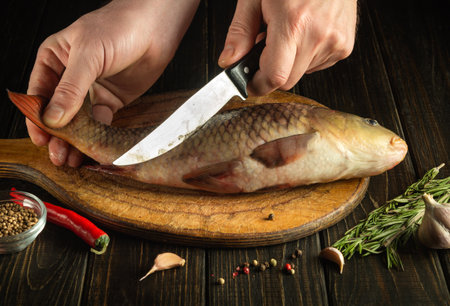 Hand of the cook with a knife cut fresh Cyprinus fish on a wooden cutting board. Home cooking of a national fish dish according to unique recipeの写真素材