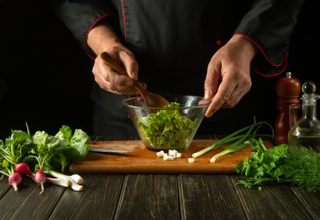 The cook mixes lettuce leaves in a bowl with a spoon. Working environment on a restaurant kitchen table. Delicious salad with fresh vegetables by the hands of the chefの写真素材