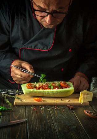 A bespectacled senior chef adds parsley to stuffed zucchini on the kitchen table. The concept of preparing a national stuffed squash or courgette for lunch. copyspace.の写真素材