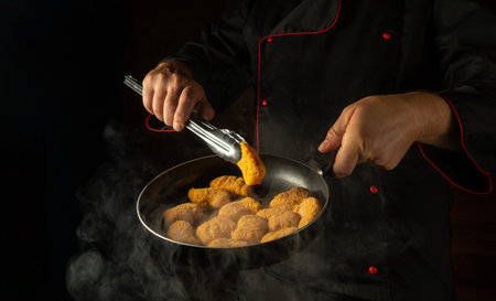 The cook prepares chicken nuggets in a frying pan. The concept of cooking nagits in the kitchen of a restaurant or hotel. Free space for advertising on a dark background.の写真素材