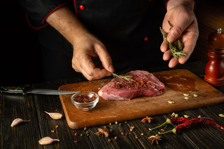 The chef adds dry aromatic rosemary to the meat steak. The process of cooking veal meat or shish kebab on the kitchen table with spices and salt.の写真素材