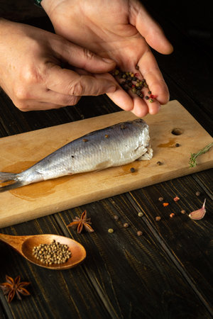 Close-up of a chef's hands adding dry pepper to Clupea fish. Concept of cooking pickled herring with spices on the kitchen tableの写真素材
