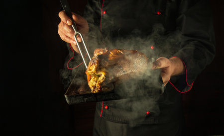 A cook bakes a chicken carcass in a restaurant kitchen. A baking sheet with a baked bird and a fork in the hands of a chef. Black space for advertising.の写真素材