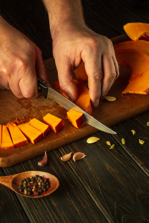 A man's hands use a knife to cut a pumpkin into small pieces on a cutting board for baking a pie. The concept of preparing a delicious national dish at home.の写真素材