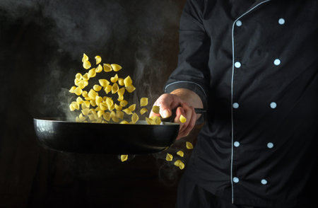 The cook throws the pasta into a hot, steamy frying pan. Italian cuisine or shell-type pasta. Place for recipe on black background.の写真素材