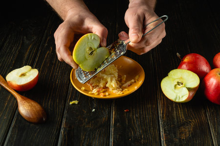 Using a hand grater to grate apples. Close-up of a man hands preparing applesauce on the kitchen table. Free advertising space.の写真素材