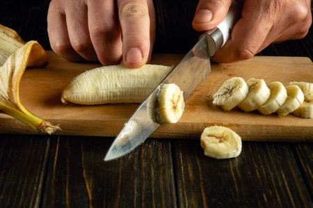 Slicing banana with knife in chef hand on kitchen wooden board for preparing fruit dessert.の写真素材