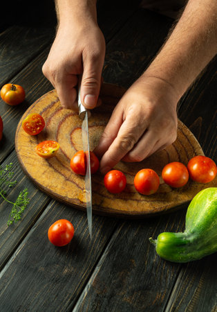 Male hands cut tomato with knife for cooking vegetable food. Cooking cherry tomato at home on kitchen table.の写真素材