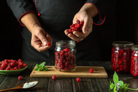 The cook fills the jar with raspberries. The process of making a sweet drink from fresh berries and sugar on the kitchen table. Preserving raspberry in a jar.の写真素材