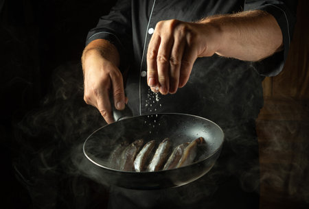 Adding salt to fish in a frying pan. Chef cooking capelin in the hotel kitchen. Concept on a dark...
