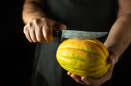 A chef tries to cut a ripe melon with a knife to prepare a dessert. Low key concept of serving melon in a restaurant. Slicing melon.の写真素材